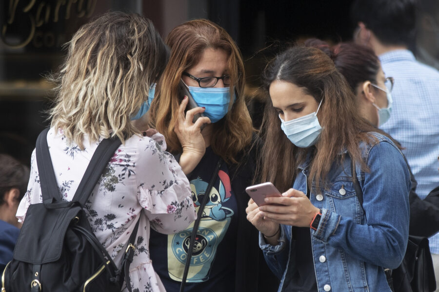 Three young women wearing masks to help prevent the spread of coronavirus stand together using their phones in Madrid, Spain, Thursday, Oct. 8, 2020. A court in Madrid has struck down a national government order that imposed a partial lockdown due to the coronavirus pandemic in the Spanish capital and its surrounding suburbs, siding with regional officials who had resisted stricter measures against one of Europe’s most worrying virus clusters. (AP Photo/Paul White)