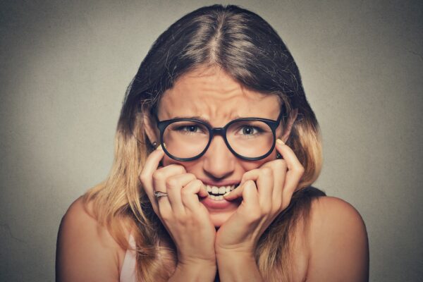 Closeup portrait nervous stressed young woman girl in glasses student biting fingernails looking anxiously craving something isolated on grey wall background. Human emotion face expression feeling