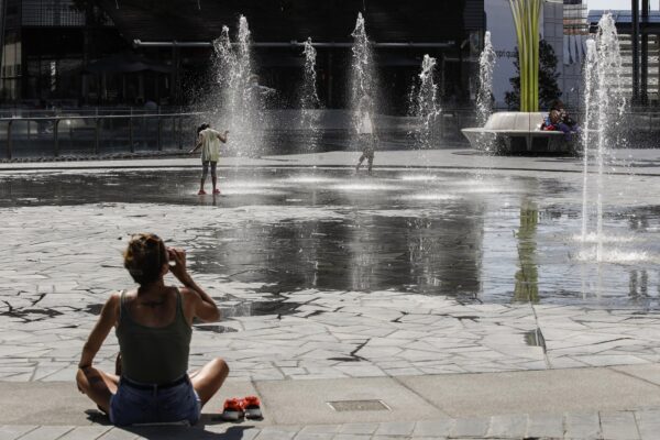 A woman drinks as children cool-off in a public fountain in Milan, Italy, Friday, July 31, 2020. The first heat wave of the summer, which arrived Thursday and will last at least until next Saturday, will reach temperatures over 34 Celsius (104 Fahrenheit). (AP Photo/Luca Bruno)