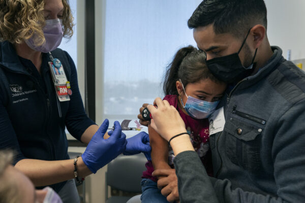 Kidney transplant patient Sophia Silvaamaya, 5, held by her father Pedro Silvaamaya, is vaccinated by nurse Kelly Vanderwende, Wednesday, Nov. 3, 2021, at Children’s National Hospital in Washington. A Buzzy is held above the injection site to minimize pain. The U.S. enters a new phase Wednesday in its COVID-19 vaccination campaign, with shots now available to millions of elementary-age children in what health officials hailed as a major breakthrough after more than 18 months of illness, hospitalizations, deaths and disrupted education. (AP Photo/Carolyn Kaster)